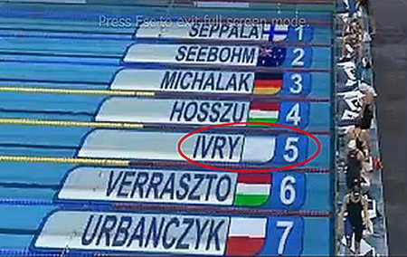 Finale im 100-Meter-Kurzbahn-Lagenschwimmen in Doha, der Hauptstadt des Katar. Bei der Live-Übertragung wie gewohnt vor dem Start die Namen der Schwimmerinnen mitsamt den Flaggen ihrer Herkunftsländer. Auf Bahn 5 fehlt die Flagge. 