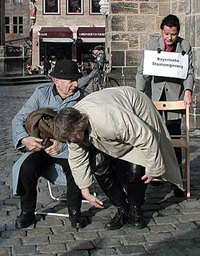 Bild: Die „Bayerische Staatsregierung“ zieht einem Obdachlosen – gespielt von einem Laiendarsteller den Stuhl unterm Hintern weg. Foto:SM/dk